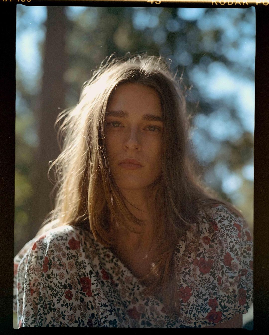 Portrait of a young adult female with long brown hair, wearing a floral print top, posing outdoors in natural light.