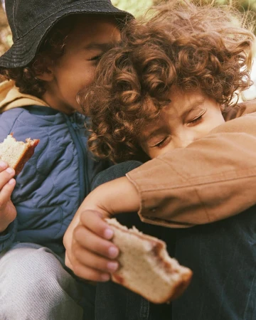 Two children with curly hair holding pieces of bread outdoors with natural light captured by Kate Moore