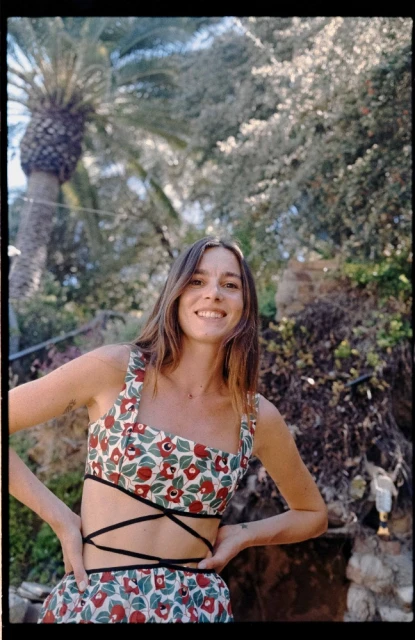 Smiling woman in floral crop top with wrap detail poses outdoors in front of palm tree and green foliage, looking at the camera.
