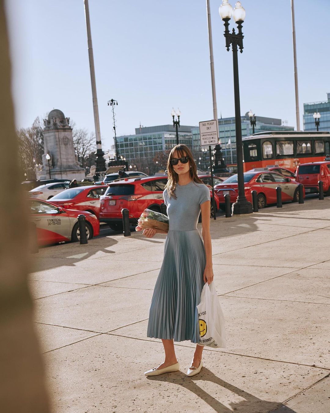 Woman wearing blue dress and sunglasses holding a grocery bag standing on a city street by parked cars, photographed by Kate Moore.