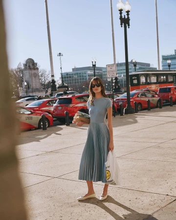 Woman wearing blue dress and sunglasses holding a grocery bag standing on a city street by parked cars, photographed by Kate Moore.