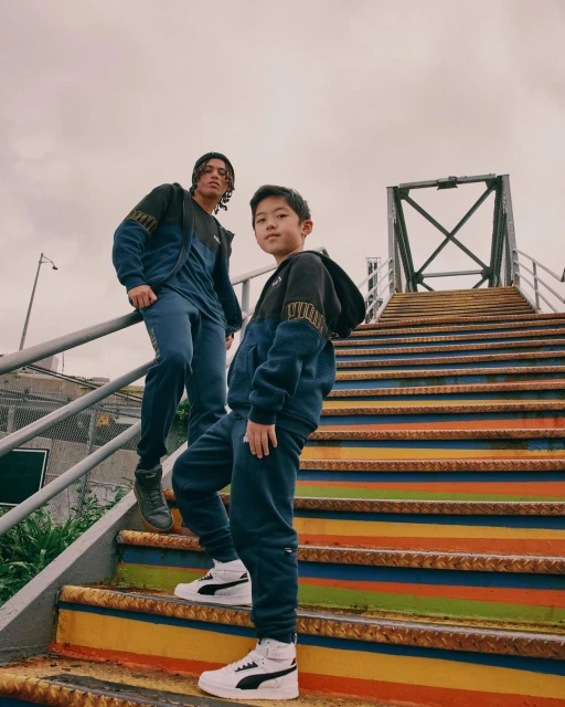Two people, one child, stand on brightly painted stairs against a cloudy sky, wearing navy and black Puma tracksuits and sneakers.