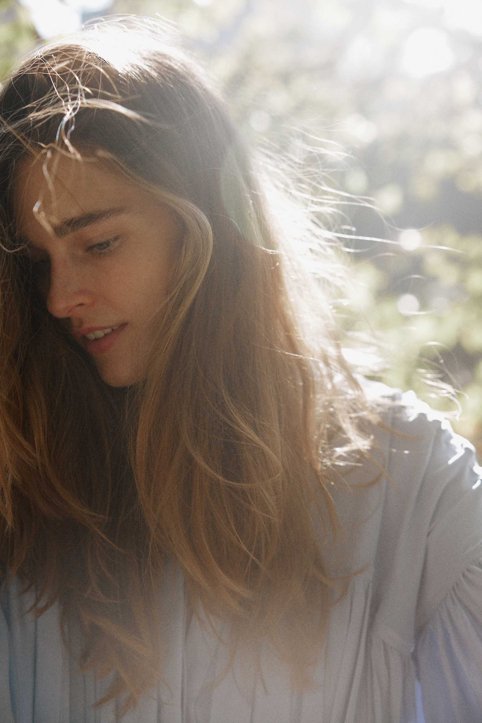 Close-up shot of a young adult woman with long brown hair, fair skin, and a light blue blouse standing outdoors with a happy expression.