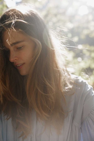 Close-up shot of a young adult woman with long brown hair, fair skin, and a light blue blouse standing outdoors with a happy expression.