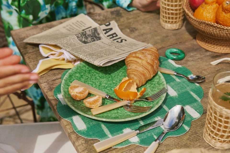 Overhead shot of a wooden table with a green plate, croissant, mandarin, silverware, wicker-covered glasses, and a newspaper titled PARIS.