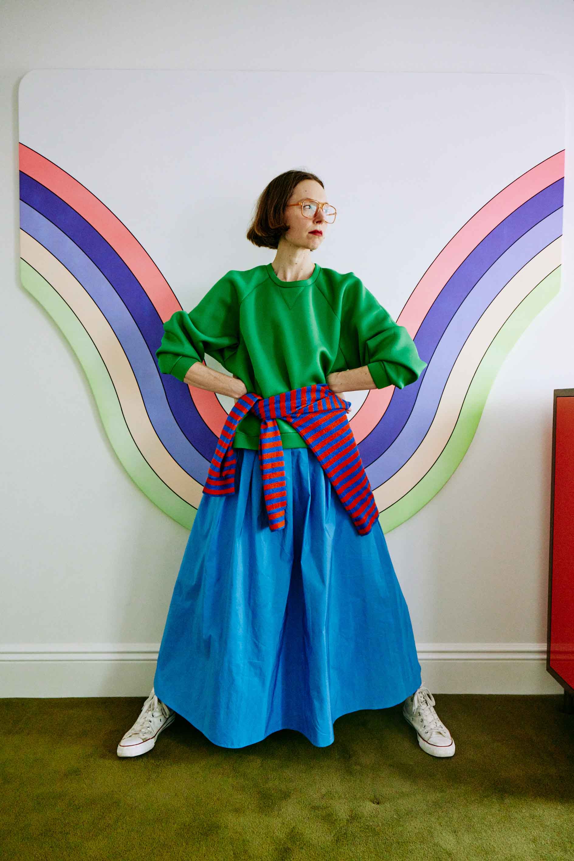 A woman poses indoors in a green shirt and blue skirt, wearing glasses and sneakers, with a colorful rainbow graphic in the background.