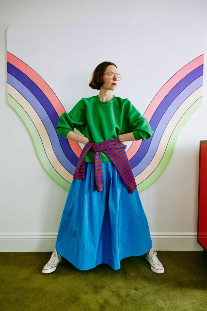 A woman poses indoors in a green shirt and blue skirt, wearing glasses and sneakers, with a colorful rainbow graphic in the background.