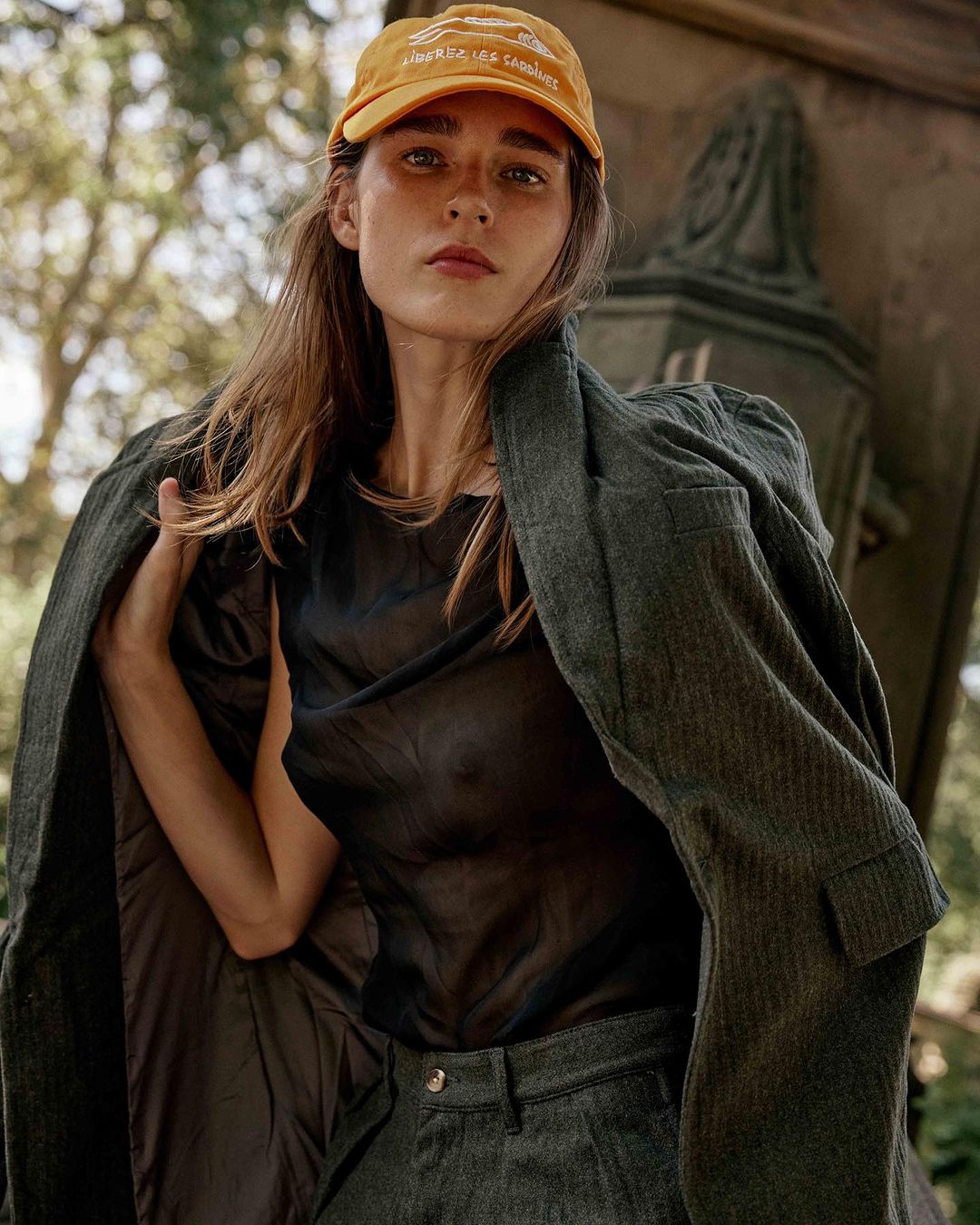 Portrait of a young adult female in a black top and blazer, wearing an orange baseball cap, posing outdoors surrounded by trees.
