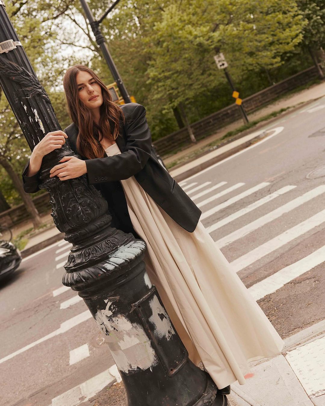 A woman in a black blazer and beige dress poses leaning on a black ornate lamppost on a crosswalk in a city street with green trees.