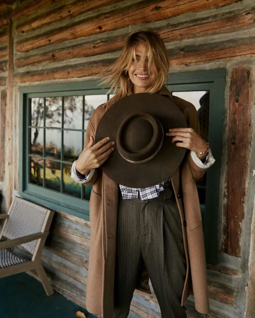 Adult woman in a tan coat and dark pants holding a brown hat on a rustic porch in front of a window in natural lighting.