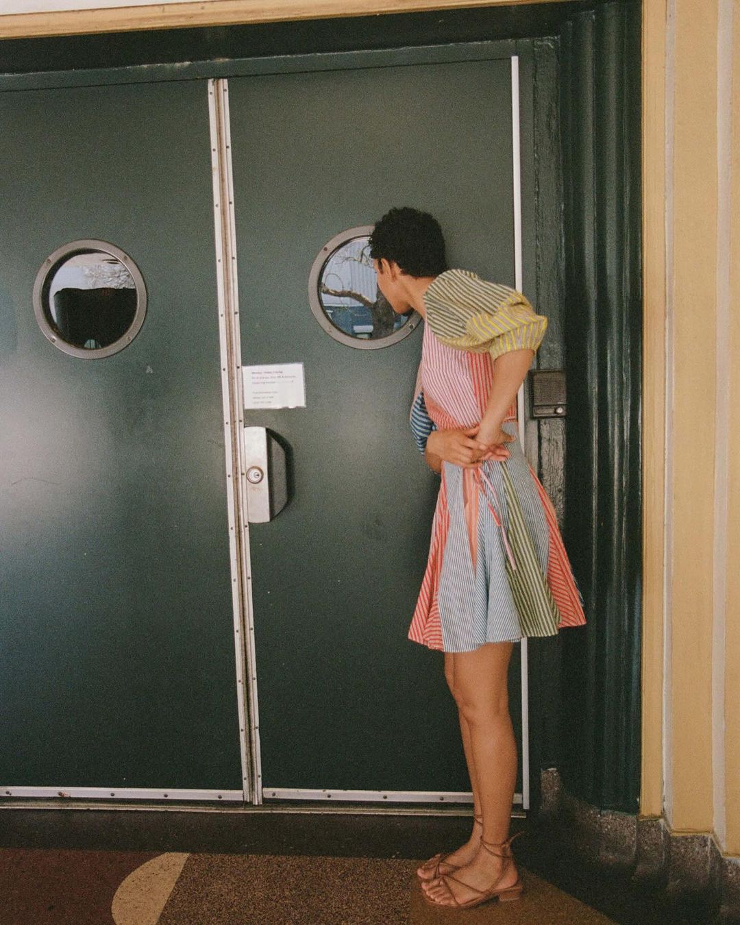 A woman in a colorful striped dress stands near a green door with porthole windows, looking into one of the round openings.
