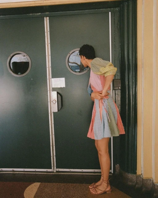 A woman in a colorful striped dress stands near a green door with porthole windows, looking into one of the round openings.