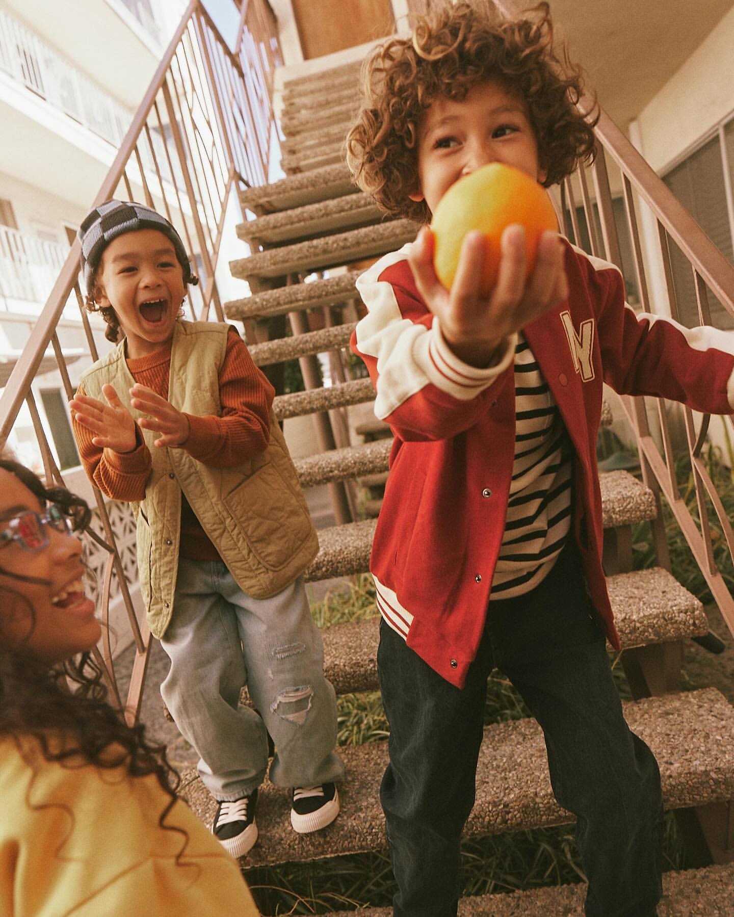 Three children, two boys and one girl, are standing outside. A boy holds an orange in his hand and the others smile at him.