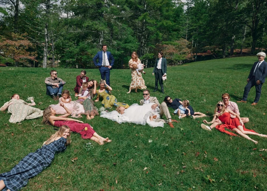 A large group of people, including a bride and groom, are lying on a grassy lawn with trees in the background, captured in a wide shot.