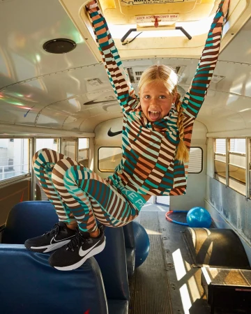 Young girl playfully hangs from the ceiling of a school bus, wearing a striped top and leggings, with sneakers, smiling at the camera.