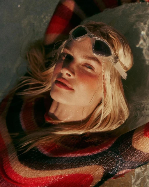 Close-up shot of a blonde woman with sunglasses lying down, wearing a multicolored, glittering top and light make-up in bright sunlight.
