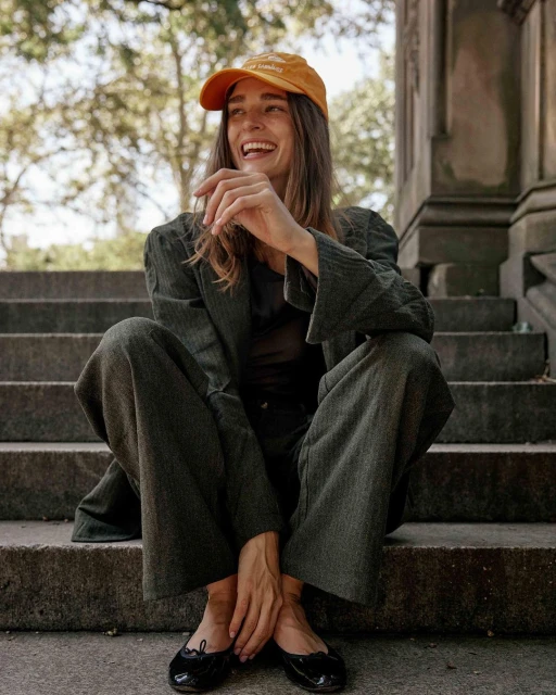 Stylish woman sits on stone steps outside, wearing a corduroy suit, orange cap, and black ballet flats, smiling in the sunlight.
