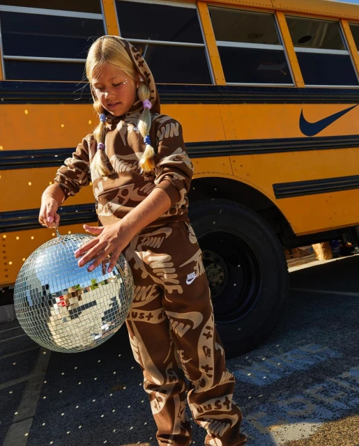 A young girl in a Nike-patterned sweatsuit poses with a disco ball in front of a yellow school bus on a sunny day.