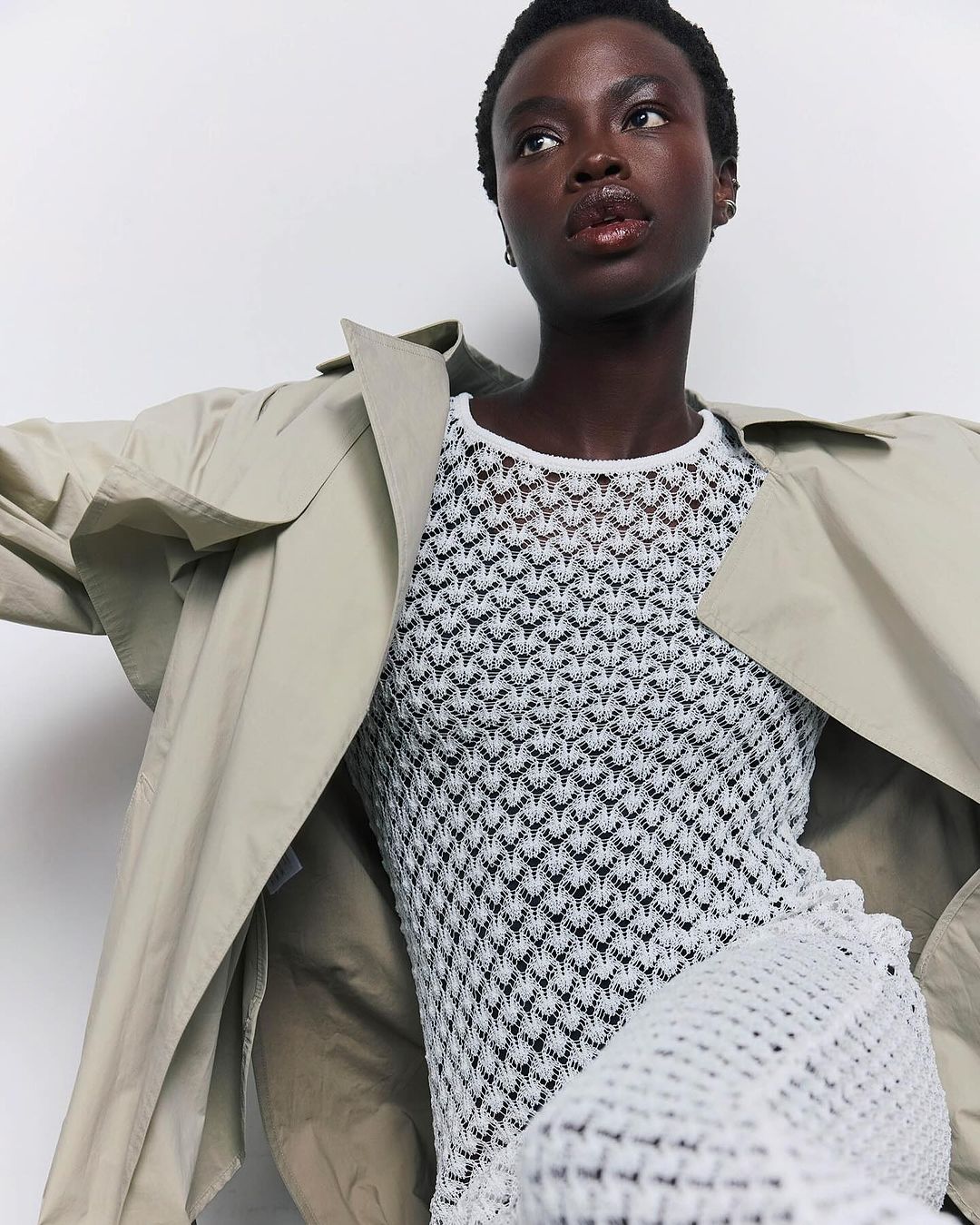 Fashionable woman with dark skin poses in a flowing trench coat over a white knit dress in front of a bright white backdrop.
