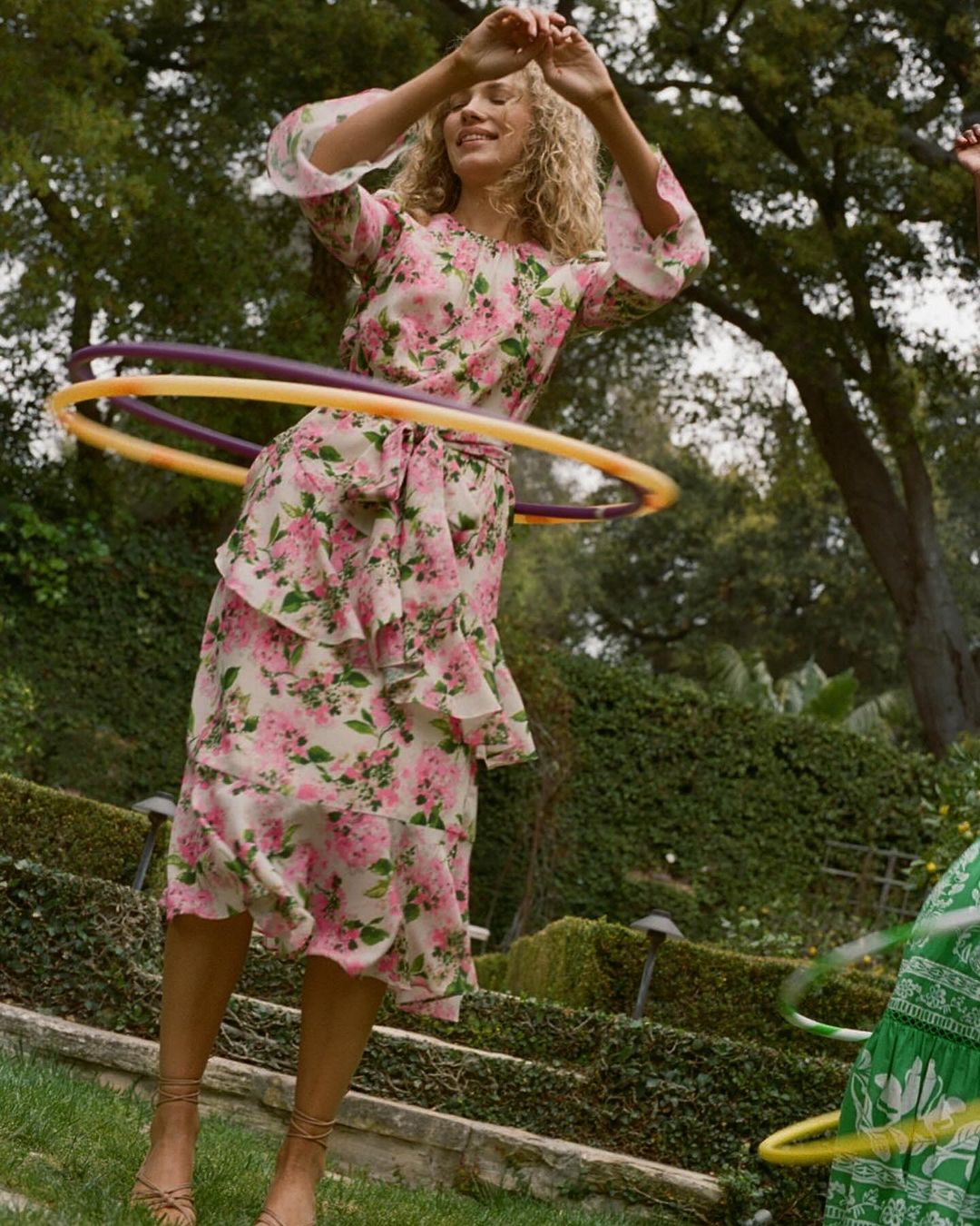 A woman outdoors wearing a floral dress and hula hooping with trees in the background on a sunny day.