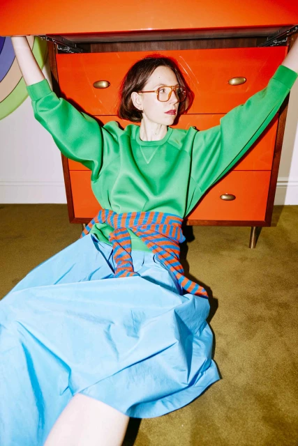 Woman in bright green top and blue skirt sits next to an orange dresser in an indoor setting with green patterned carpet.