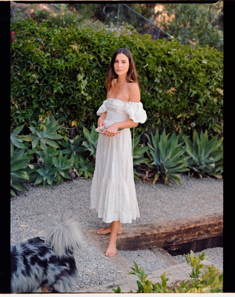 Full-length shot of a brunette woman in a white dress standing in a yard with a dog, surrounded by plants and greenery on a sunny day.