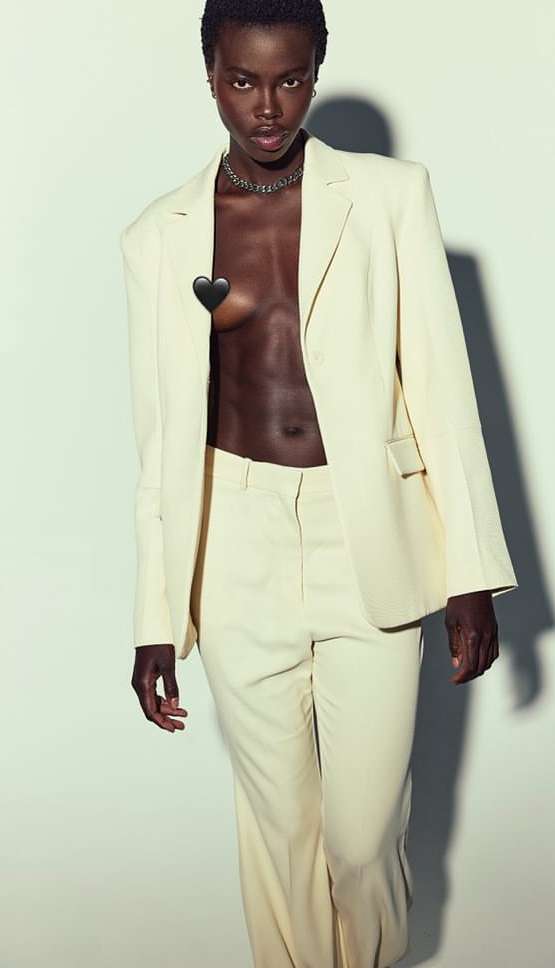 Elegant dark-skinned woman poses in a light yellow suit with silver chain necklace, captured in a studio setting with soft lighting.