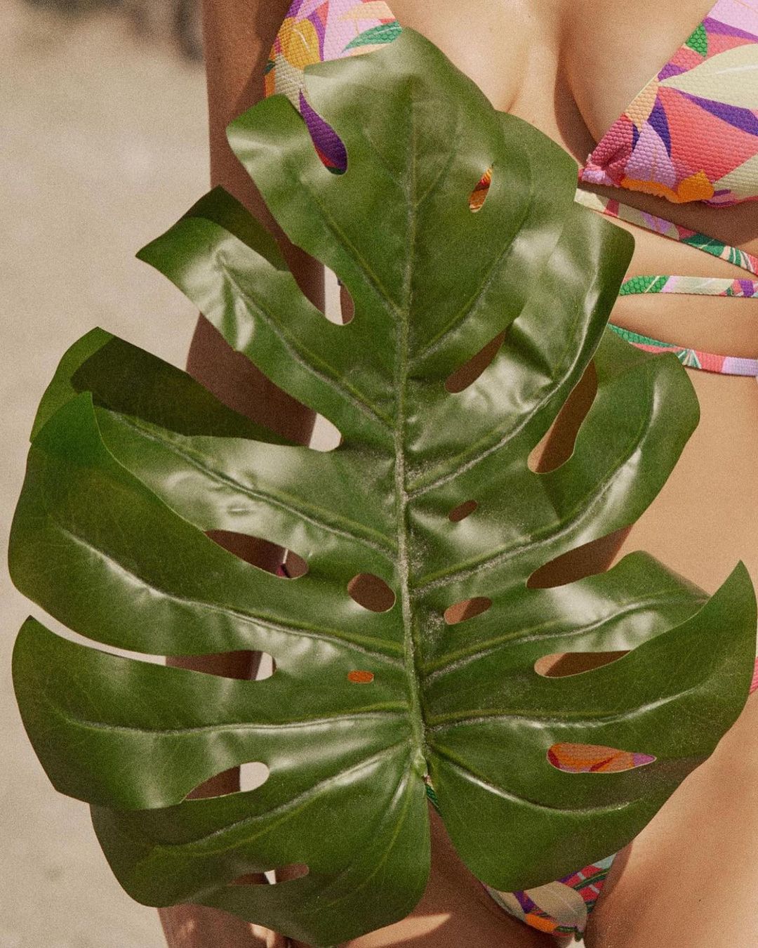 Woman in a bikini partially obscured by a green monstera leaf, standing outdoors with bright natural lighting and a tan sandy background.