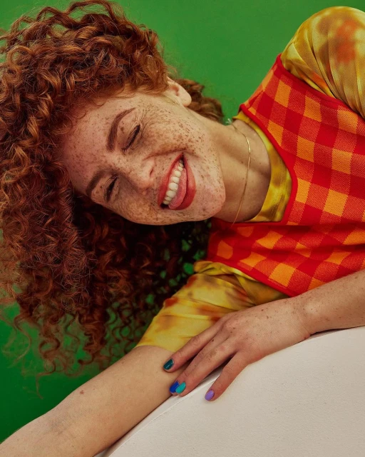 Close-up studio shot of a happy adult woman with red curly hair and freckles wearing a red-and-yellow patterned top against a green background.