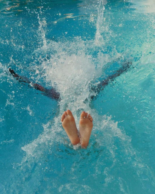 Person dives into a turquoise-blue pool, legs and feet visible above the waterline amid splashing water in an indoor swimming pool.