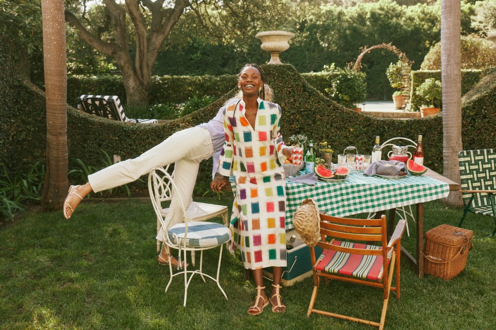 A cheerful Black woman in a colorful dress stands on a lawn with a picnic table, garden and another person with legs on a chair.