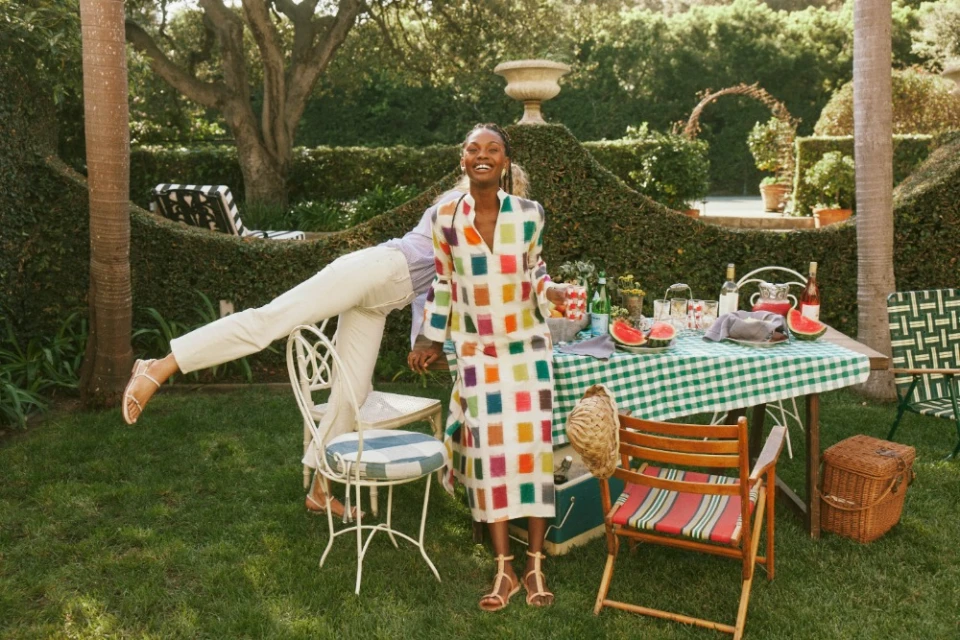 A cheerful Black woman in a colorful dress stands on a lawn with a picnic table, garden and another person with legs on a chair.
