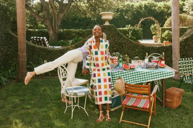 A cheerful Black woman in a colorful dress stands on a lawn with a picnic table, garden and another person with legs on a chair.