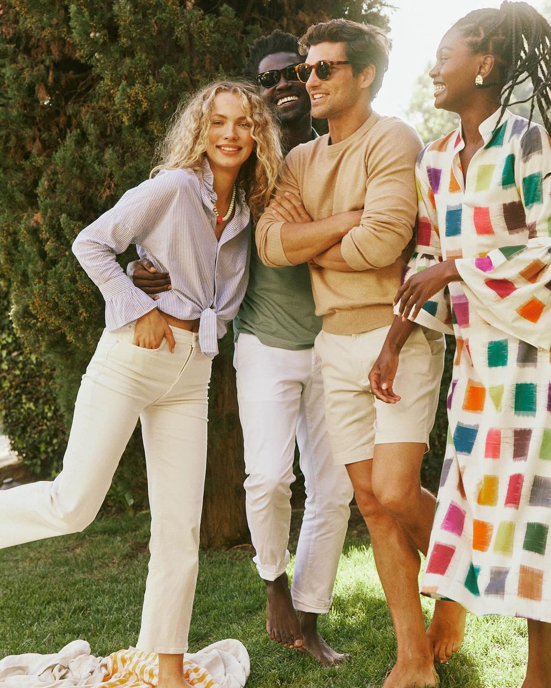 Group of four adults outdoors on grass. Two women, one in colorful dress, one in striped shirt and white pants. Two men in casual wear.