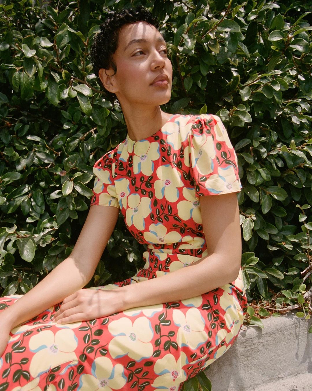A woman with short dark hair poses outdoors, wearing a floral print dress in red, yellow, and white, with a green leafy background.