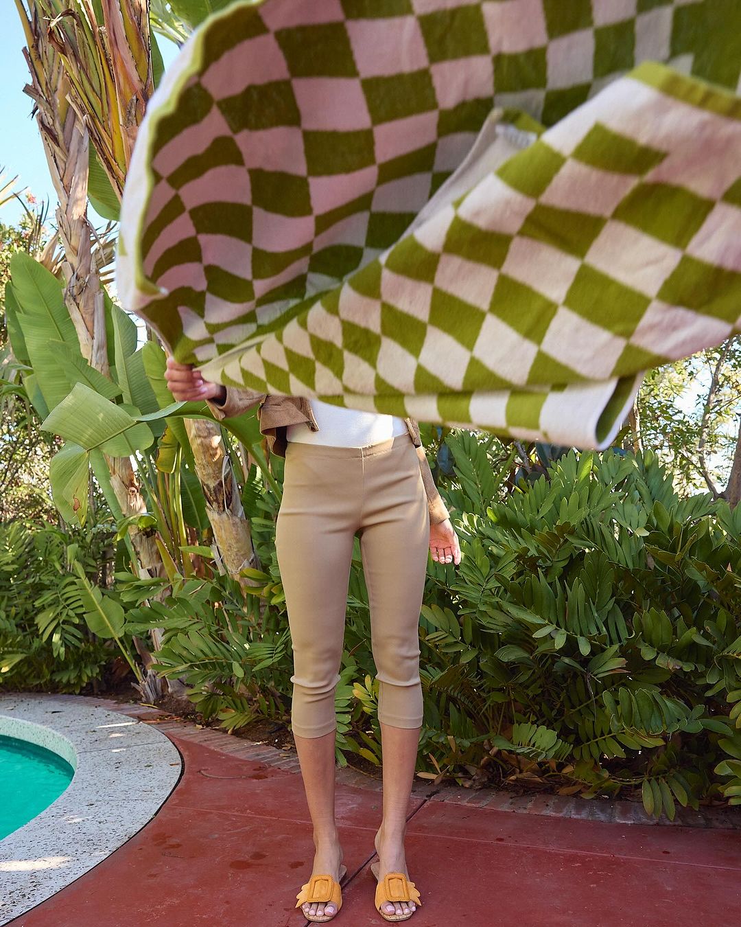Woman stands by a pool holding a green and white checkered towel, wearing capri pants, a jacket, and sandals in an outdoor setting.