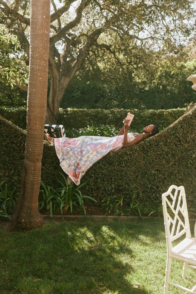 Woman in patterned dress relaxes on outdoor hedge hammock reading a book, with a palm tree and green lawn, in the daytime.