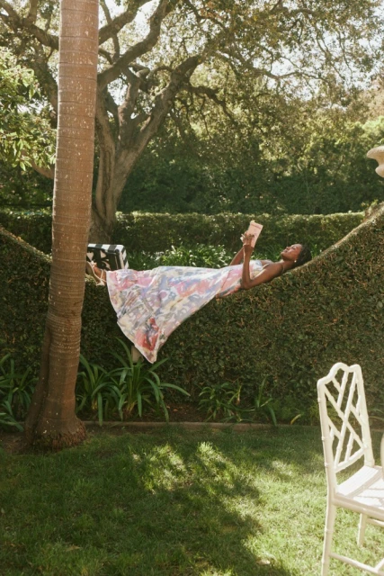 Woman in patterned dress relaxes on outdoor hedge hammock reading a book, with a palm tree and green lawn, in the daytime.