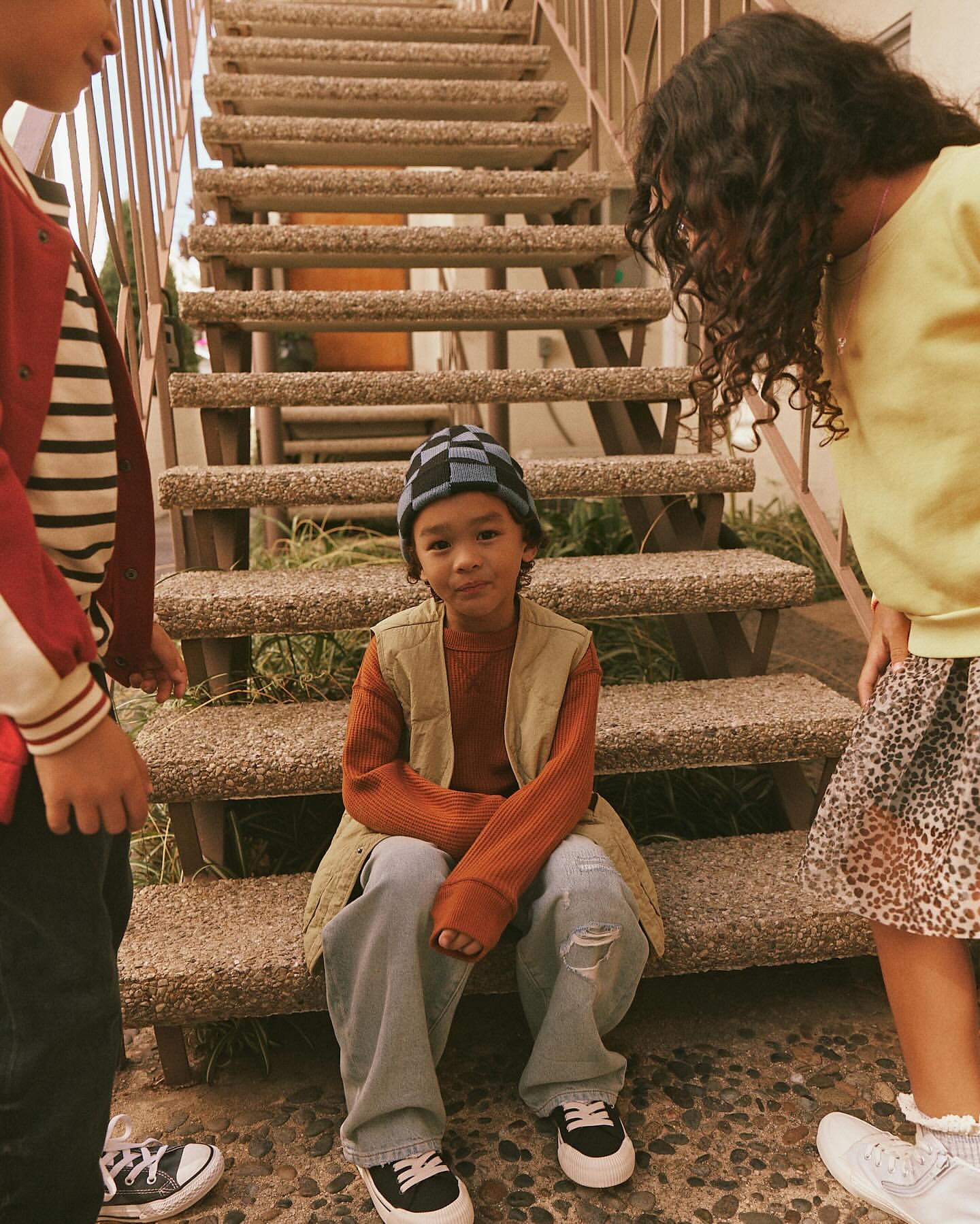 Child in vest sits on concrete steps, with other children standing nearby wearing casual streetwear fashion, outdoor shot, medium framing.
