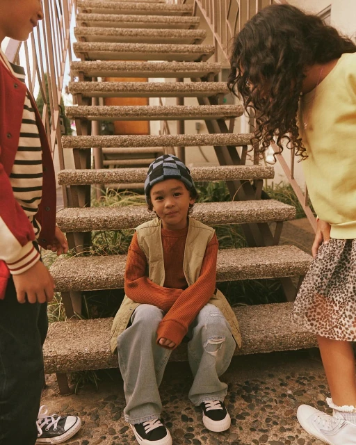 Child in vest sits on concrete steps, with other children standing nearby wearing casual streetwear fashion, outdoor shot, medium framing.