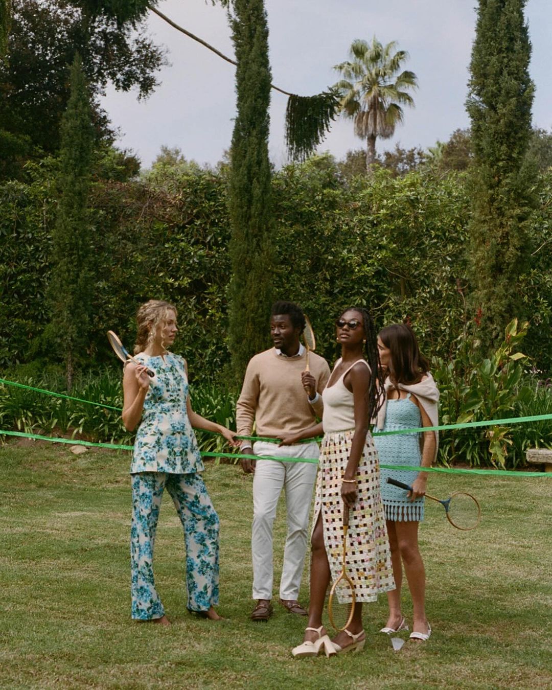 Four adults on a lawn playing lawn tennis with vintage rackets. Two women in dresses, one in a pantsuit, and one man in tan sweater.