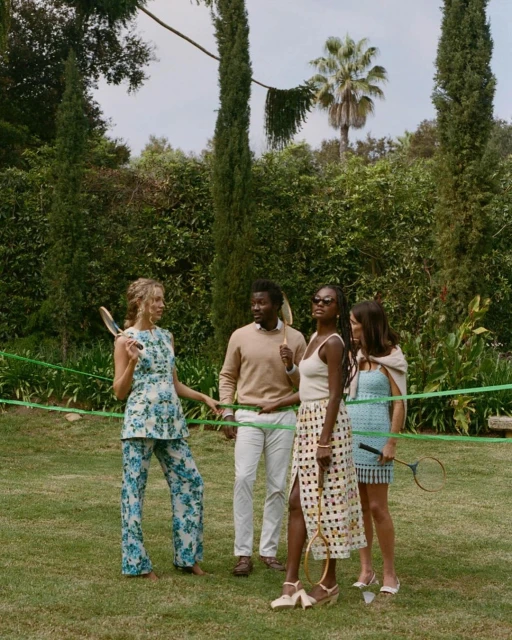 Four adults on a lawn playing lawn tennis with vintage rackets. Two women in dresses, one in a pantsuit, and one man in tan sweater.