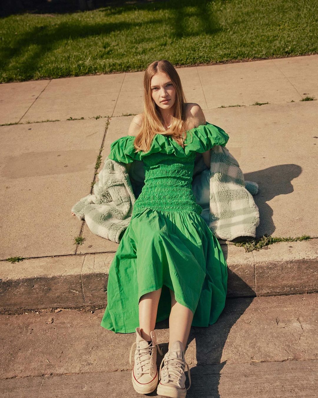 Young woman poses outdoors wearing a green off-the-shoulder dress and tan sneakers, seated on concrete with a jacket beside her.