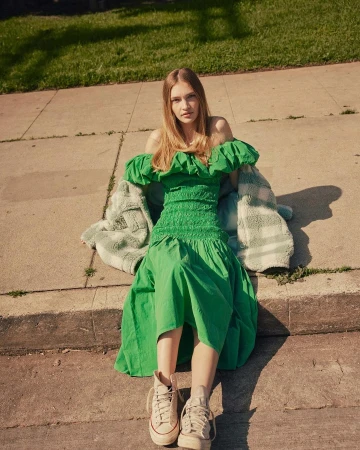 Young woman poses outdoors wearing a green off-the-shoulder dress and tan sneakers, seated on concrete with a jacket beside her.