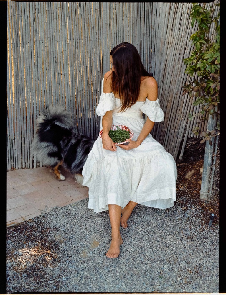 A woman in a white dress sits in a garden setting next to her dog. She is holding a bowl of wheatgrass.
