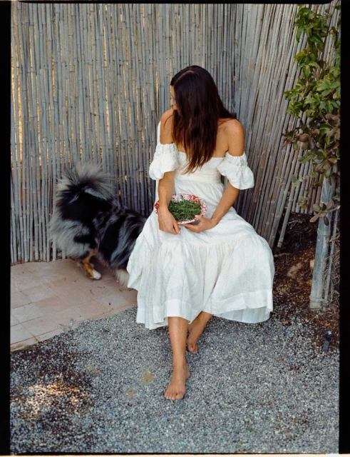 A woman in a white dress sits in a garden setting next to her dog. She is holding a bowl of wheatgrass.