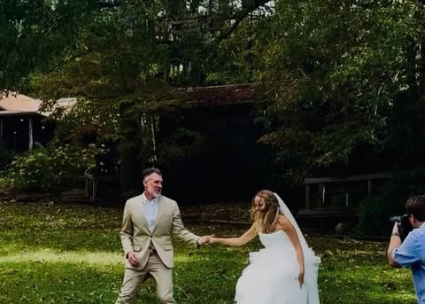 Outdoor wedding photo featuring a bride in a white dress and groom in a tan suit holding hands on a lawn, with a photographer present.