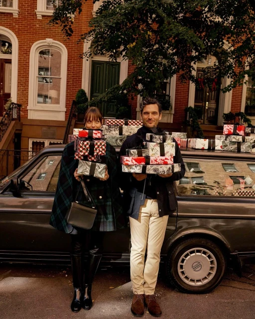 A man and woman stand smiling on a city street in front of a car, each holding stacks of wrapped gift boxes in their arms.