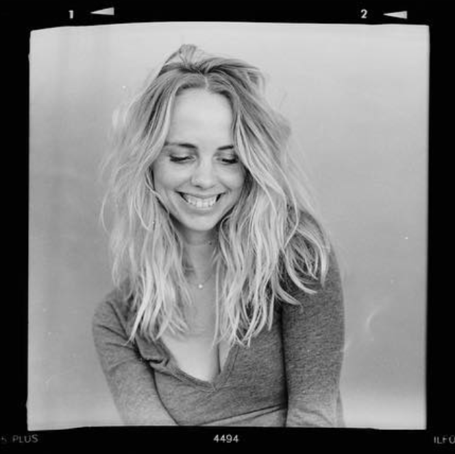 Monochrome portrait of a smiling woman with long wavy hair, wearing a casual top. The image has a vintage film border.