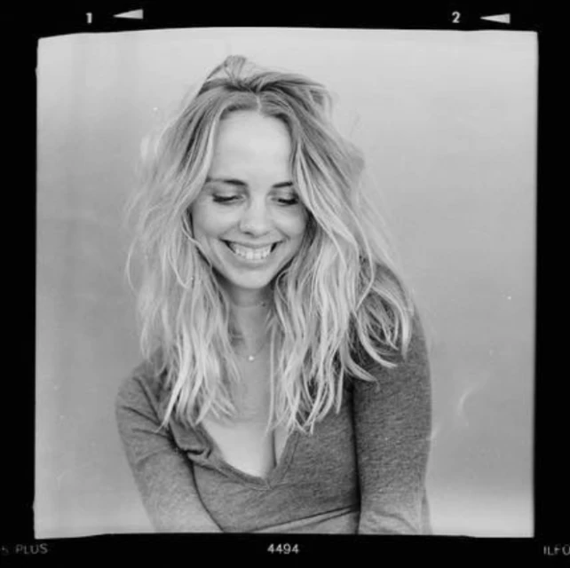 Monochrome portrait of a smiling woman with long wavy hair, wearing a casual top. The image has a vintage film border.